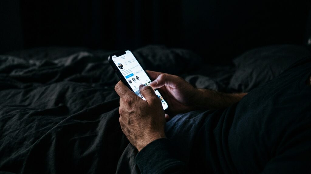 Manos de un hombre usando un teléfono inteligente brillante en la oscuridad de su cama, ejemplificando el impacto de la luz azul en el descanso.